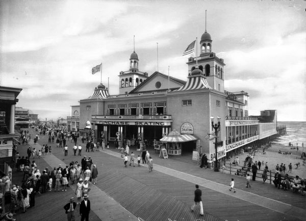 Crowds walk along the boardwalk at Funchase amusement pier.  A building with flags flying from its turrets features a sign that reads "Funchase Skating". The beach is visible to the right and other shops and buildings stand to the left.