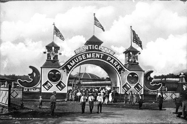 People stand at the gate to the ten-acre Monticello Amusement Park. The arched entrance is decorated with various shapes, windmills, towers, and flags. Fences stand on either side of the entrance and other buildings can be seen in the background.