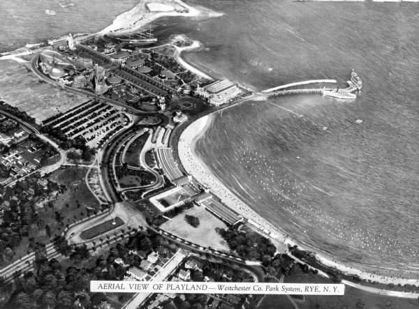 Bird's eye view of Playland, part of the Westchester Co. Park System. View features the park, the shoreline, the ocean, and several ships docked along the shore.