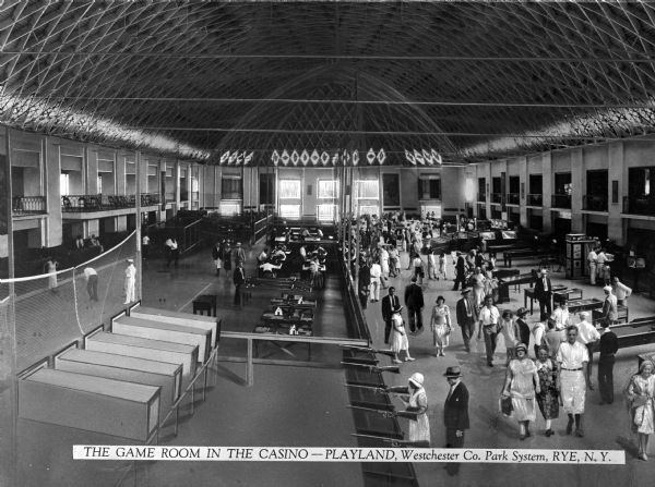 Interior view of the casino at Playland, part of the Westchester Co. Park System. Crowds of people gather inside the room, which features an elaborate ceiling, windows, amusements, and balconies on either side.