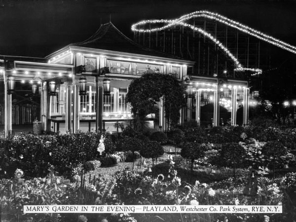 View of a garden at Playland, a Westchester Co. Park. The nighttime photograph features an illuminated building and roller coaster in the background.