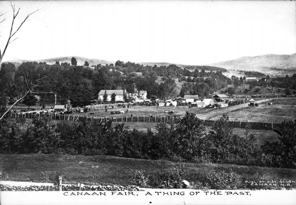 Distant view of a fair being held in a field.  Animals, buildings, fences, a road, trees, and hills surround the field.