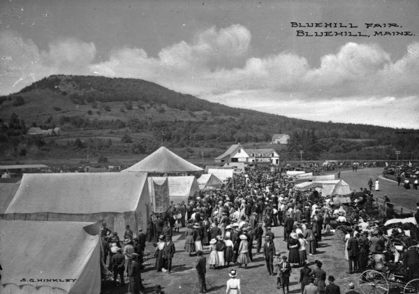 People walking around tents at the Bluehill Fair.  A road stands to the right and hills are visible in the background.