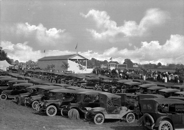 Automobiles park beside several buildings on the fairgrounds.  A crowd of people gathers in the background near a grandstand.