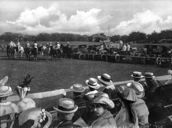 Spectators watch a horse show from a grandstand while others stand near automobiles in the background.  A building and a flag can be seen nearby.