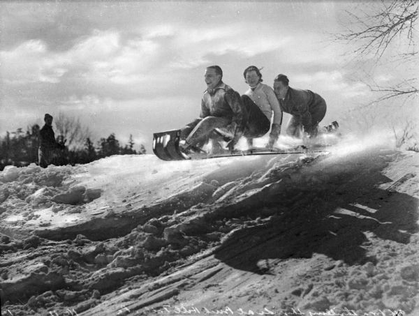 Two men and a woman sit on a toboggan in midair while riding down a snow-covered hill.