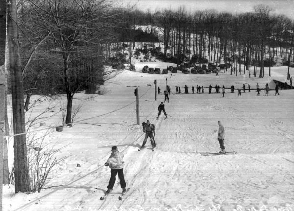 Several skiers are pulled up a snow-covered hill by a ski tow while several others wait in line and another skier looks on from the hill.