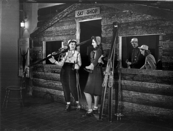 Two women with ski equipment stand in the entrance of a building with a sign overhead reading, "Ski Shop." A man and a woman stand inside the shop looking at skis.