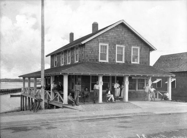 A road passes in front of the Fish and Gun Club, a two-story building with people sitting and standing on the porch.  Another dwelling stands beside it and a body of water is in the background.