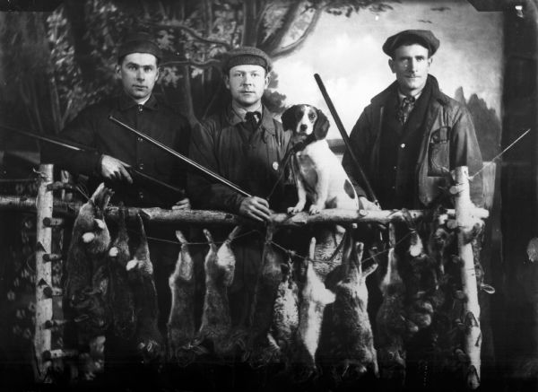 Three hunters holding rifles display their rabbits hanging from a wooden fence.  A dog also sits on the fence.