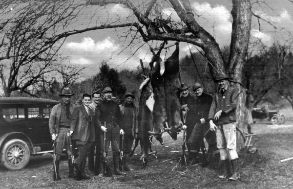 A hunting group stands around three deer strung from a tree along the side of a road.  Automobiles are parked in the background.