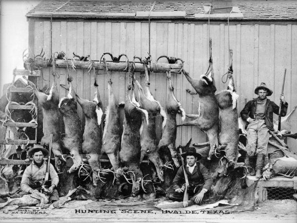 Three hunters pose with deer and antlers strung along the side of a building.