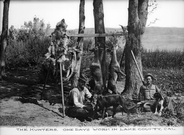 Two hunters and their dogs sit in front of three hanging deer and an animal fur strung from a wooden beam. There are trees and hills in the background.