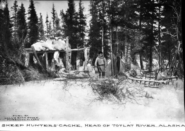 View of Sheep Hunters' Cache at the head of the Totlat River in Alaska featuring a hunter in a wooded campsite surrounded by trees and snow.