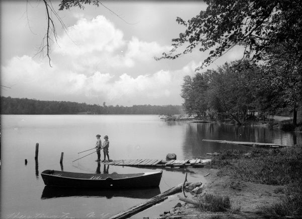 Two children stand at the end of a pier while fishing in a lake. A boat is in the forefront of the image.
