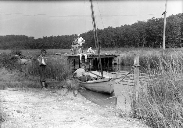 Several people stand and fish off of a pier while a boy sits in a sailboat and another stands on shore. Trees and power lines are visible in the background.