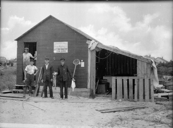 Four fishermen for hire stand with fish and fishing equipment in front of their store. The store features a doorway entrance with an open storage area.