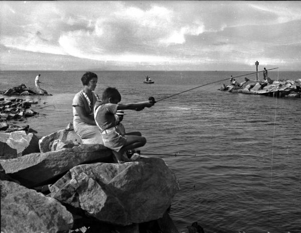 A woman and a boy fish from a rock outcrop in the foreground of the photograph, while several other people fish from a boat and rocks behind them.