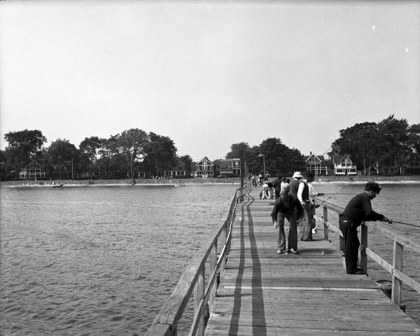 Men fishing from a wooden bridge in front of a shoreline lined with dwellings and trees.