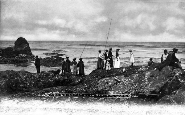 A group of men, women, and children fishing from a rock outcrop along the pacific coast. Caption reads: "Fishing Rocks, Near Seaview, Wash."