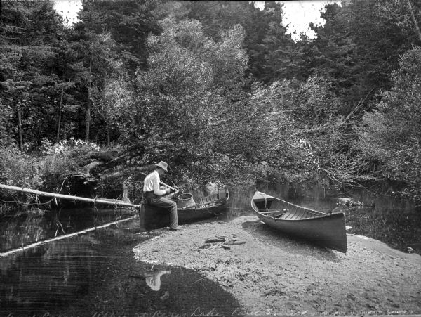 A man sits in a floating canoe filled with fishing equipment while another canoe rests on a nearby shore.