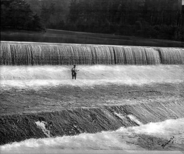 A man fishes for trout at the foot of a waterfall.