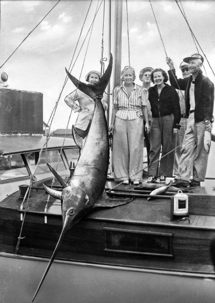 Fishermen and women pose with a swordfish tied to a mast aboard a boat.