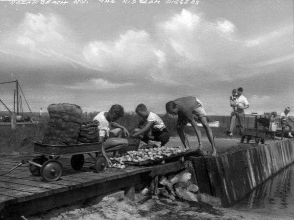 Three young men digging for clams along a pier.  A man and children are visible in the background.