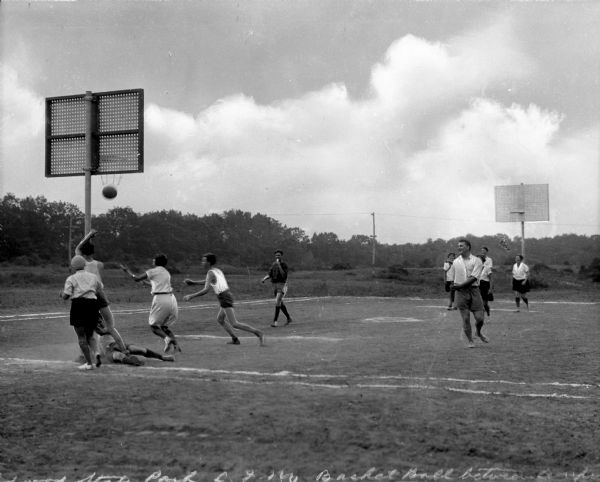 Young male and female campers in casual dress play basketball on a grass court with trees and power lines in the background.