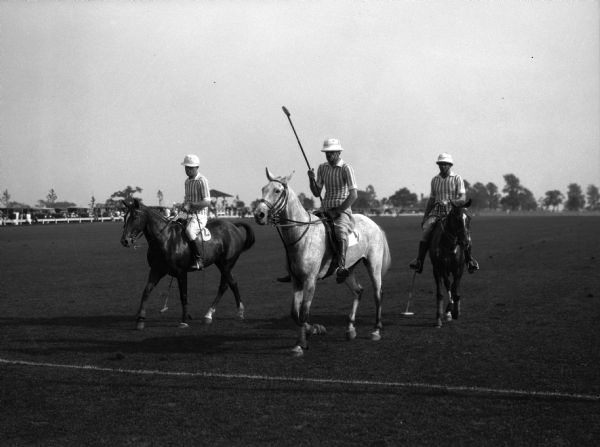 Three men on horseback wear team uniforms on a grass polo field.  A fence and automobiles are visible in the background at left.