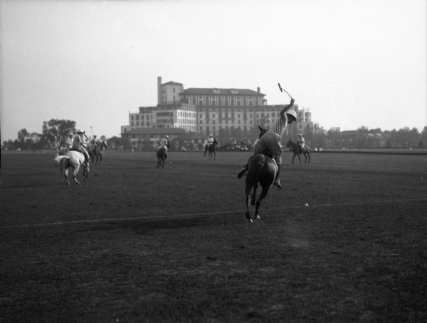 Polo players on horseback wear team uniforms while playing on a grass field.  A multi-storied building stands in the background.