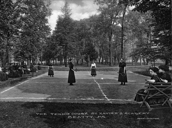 Women wear uniforms while playing tennis at St. Xavier's Academy.  Other women sit on wooden benches alongside the court and trees surround the area.