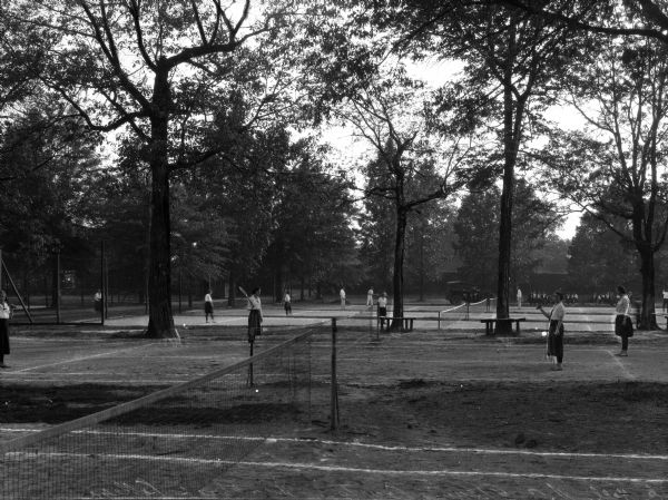 Students in uniforms play tennis on grass tennis courts surrounded by trees at Winthrop College. Students sit next to an automobile in the left background.