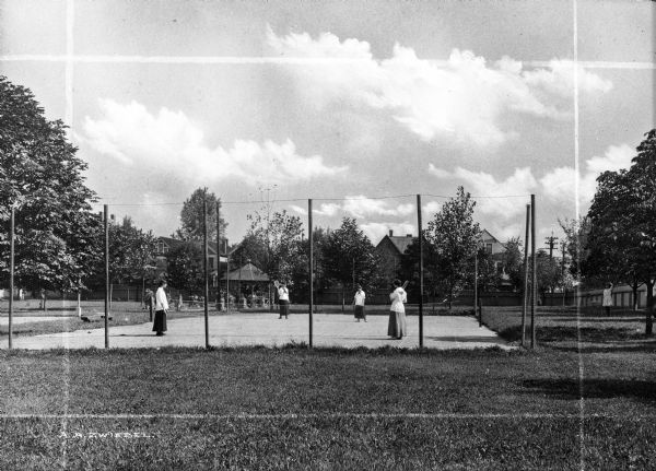 Four uniformed female students play tennis on a fenced-in tennis court at St. Ann's school. The court is surrounded by trees, a pavilion, and  buildings are visible in the background.