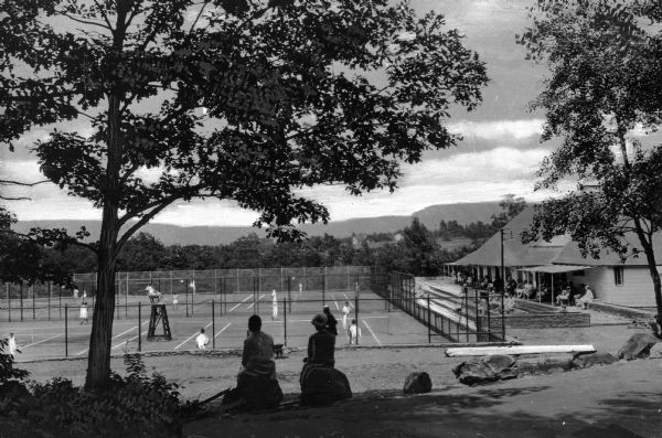 People use outdoor tennis courts while others watch from beneath the shade of a clubhouse awning.  Others in the foreground also look on while sitting on rocks beneath trees.