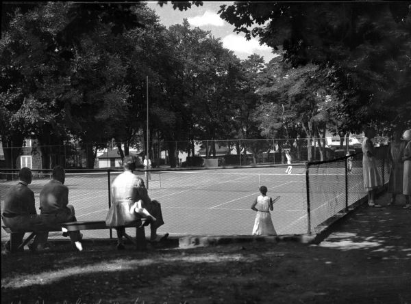 Spectators sit and stand to watch people play tennis on a fenced-in tennis court at Shelter Island Height. The court is surrounded by trees and several buildings can be seen in the background.