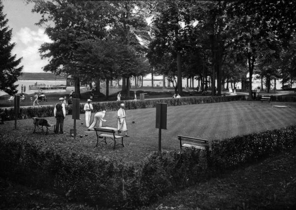 Several people use a lawn bowling court at Chautaugna Lake.  The court is surrounded by a border of shrubbery, and a dock on the lake is visible through trees in the background.
