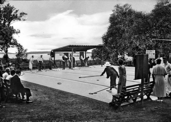 Several people play shuffleboard on an outdoor court at Lake Chautaugna.  Spectators sit on benches and stand around the court to watch the games.