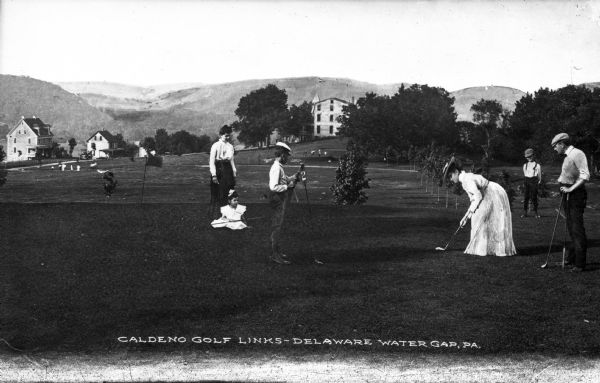 Men and women play golf at Caldeno Golf Links.  Several buildings and hills are visible in the background.