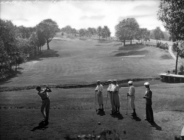 Five men look on as another man takes a swing at a golf ball on Buck Hill Falls golf course.