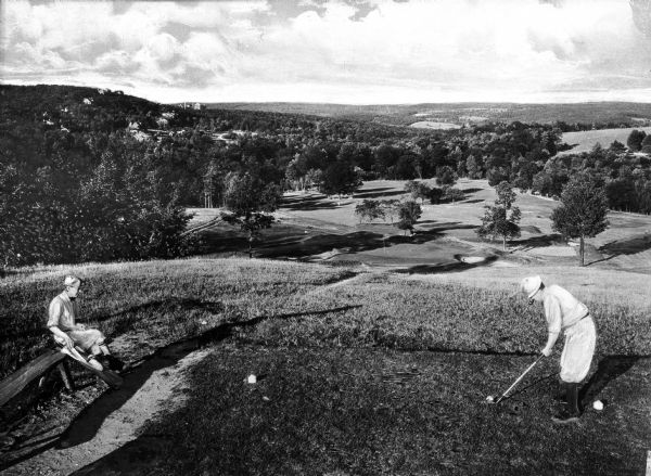 A golfer tees off from atop a hill on a golf course as another man looks on from a nearby wooden bench.