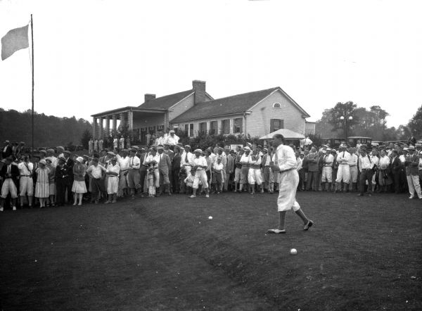 A golfer walks across a course at Lake Chautauqua while a group of spectators looks on.  A building, possibly the clubhouse, and a flag stand in the background.