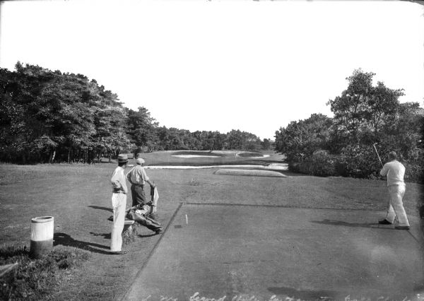 Three golfers play the second hole on South Hampton Golf Club.  Sand traps are visible between trees in the background.