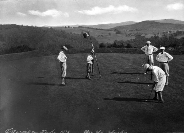 A man putts on a hilltop hole of a golf course while four others look on.