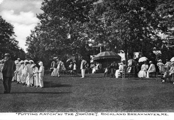 Spectators stand beneath the shade of trees and umbrellas while watching competitors in the Samoset Resort outdoor putting contest.
