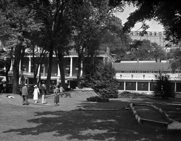 A group of people play on a hole at the Bedford Springs Hotel Miniature Golf Course.  The hotel stands behind them and has multiple stories, some with balconies.
