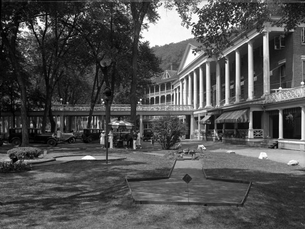 Several people stand near an umbrella-covered table outside Bedford Springs Hotel and Miniature Golf Course. The golf course stands in the foreground and the multi-storied hotel is visible in the background.