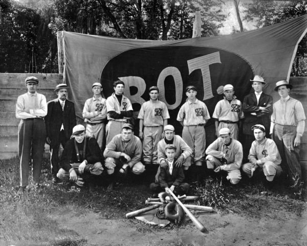 A baseball team poses for a group portrait in front of a banner reading, "B.O.T." A pile of baseball bats, balls, and mitts lies in front of the group.
