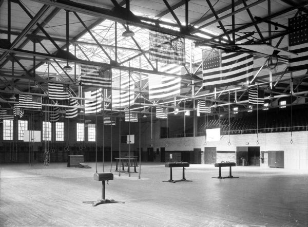 American flags hang from the ceiling in the Kent State College Wells Gymnasium. The gym holds a variety of fitness equipment including rings, parallel bars, climbing ropes, and pommel horses.