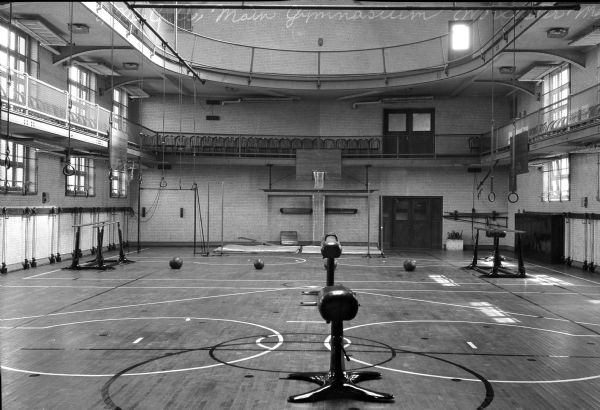 The Y.M.C.A. Main Gymnasium features athletic equipment including pummel horses, parallel bars, rings, a basketball hoop, and several balls.  The room has an upper and a lower balcony, the lower lined with folding chairs.
